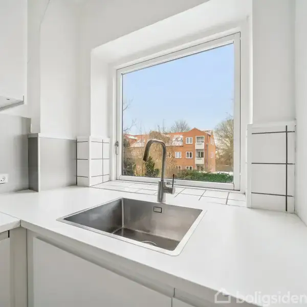 En køkkenvask med en høj hane er monteret i en hvid bordplade. Bagved ses et stort vindue med udsigt til murstensbygninger og træer udenfor.