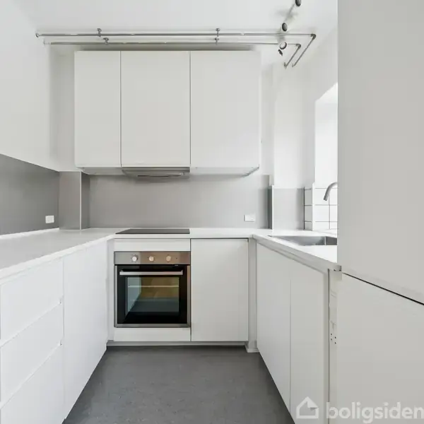Et moderne, hvidt køkken med indbyggede skabe og integreret ovn. Det er i en L-formet opsætning med grå vægge og spotlight-lys. En hvid vask ses på højre side.