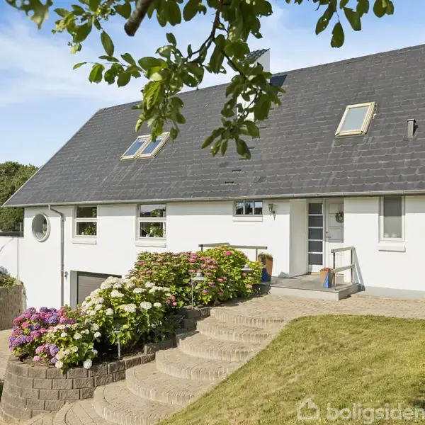Hvidt hus med skråtag og flere tagvinduer står i en velplejet have. En trappe omgivet af blomsterbede fører op til hoveddøren. Græsplæne og træer omkranser huset.