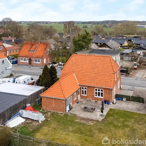Et rødt murstenshus med en karakteristisk orange tagkonstruktion står i en villakvarter. Flere lignende huse kan ses i baggrunden, omgivet af veje og grønne områder.