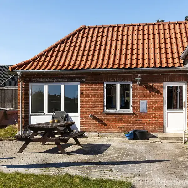 Rødstenshus med rødt tegltag, havebord og parasol på terrasse. Græsplæne og hegn omkranser området. Døre og vinduer i hvid ramme leder ind til huset. Blå blomsterkrukke ved siden af døren.