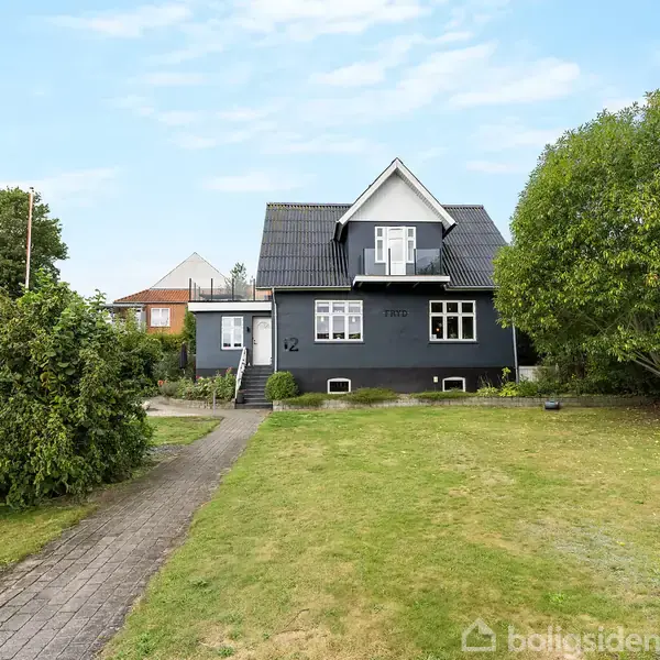 Hus med sort tag og facade står foran en grøn græsplæne med træer og buske. Teksten "1939" og "12" er synlig på facaden.