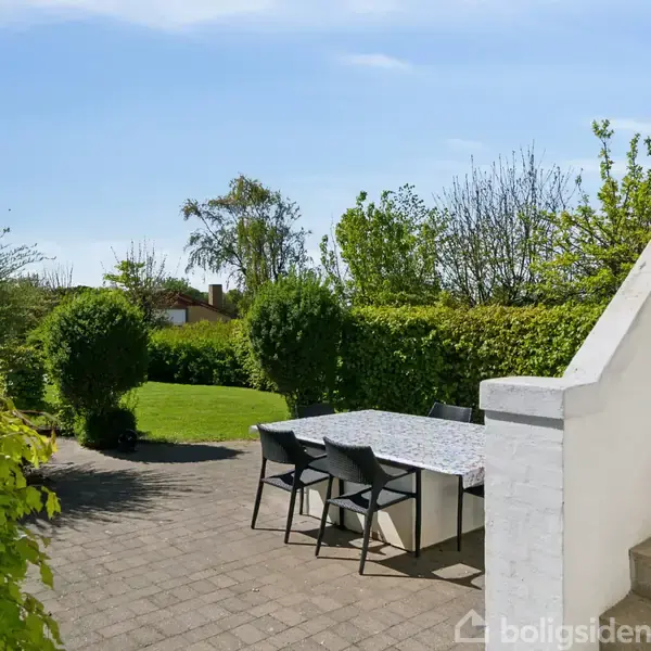En udendørs terrasse med et kvadratisk bord og fire sorte stole placeret på flisebelagt gulv. Haven er frodig med grønne buske og græsplæne, og der er en murtrappe opad.