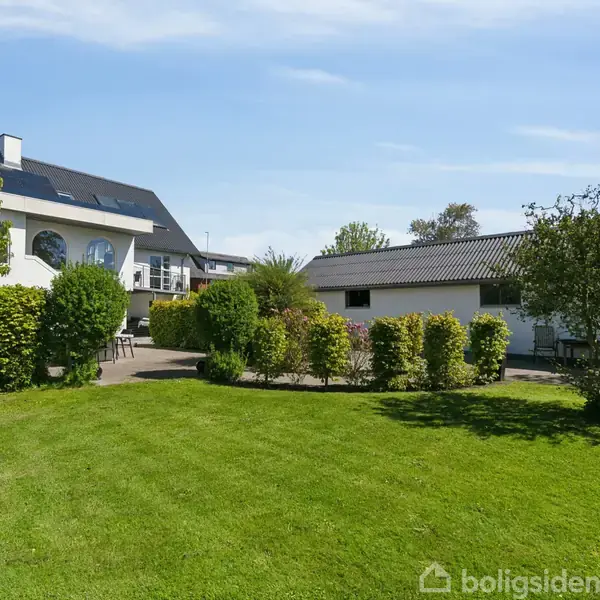 Hus med solpaneler på taget står stille. Haven har grønne buske og en velplejet græsplæne. Et mindre udhus ses i baggrunden under en klar blå himmel.