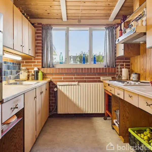 Køkken med træskabe og murstensvægge. Køkkengrej er placeret på bordpladerne. Sollys strømmer ind gennem et vindue over en radiator. En skål med æbler står på gulvet.