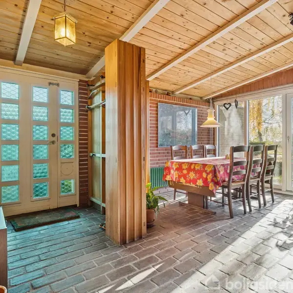 En spisestue med bord dækket af rødt mønstret dug. Seks stole er omkring bordet. Området er aflangt med glasdør til haven. Murstensvægge og træloft præger rummet.