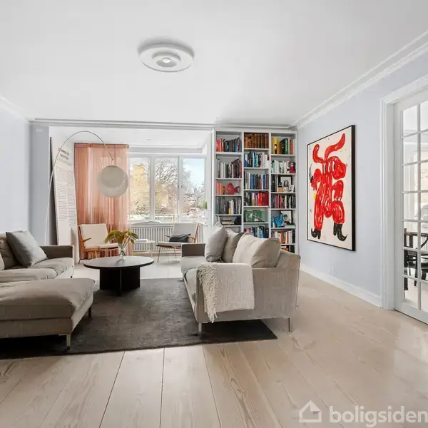 Sofaer om bord i stue med lyse trægulve og store farverige kunstværker på væggene; hylder fyldt med bøger og glasdøre fører til næste rum.