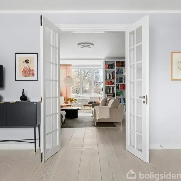 Glasdøre åbner til en moderne stue med en beige sofa, reol fyldt med bøger og stor vindue med gardiner. Sidebord med sort lampe og kunstværk hænger på væggen.