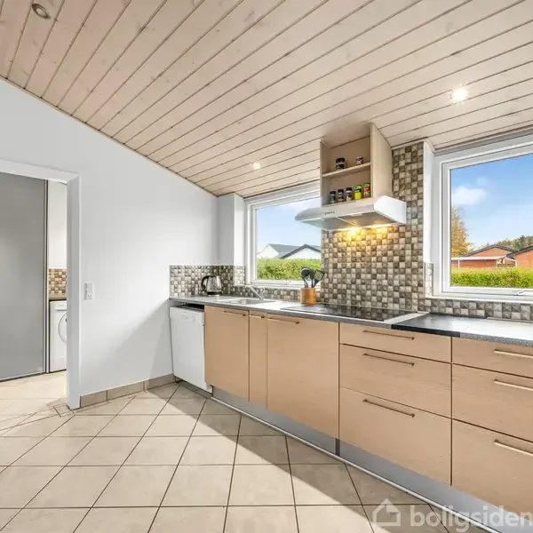 Køkkenet er moderne med lyse træskabe og stålhåndtag, udstyret med opvaskemaskine, vask og kogeplader. Væggene er mosaikfliser, og store vinduer giver udsigt til en have med blå himmel.