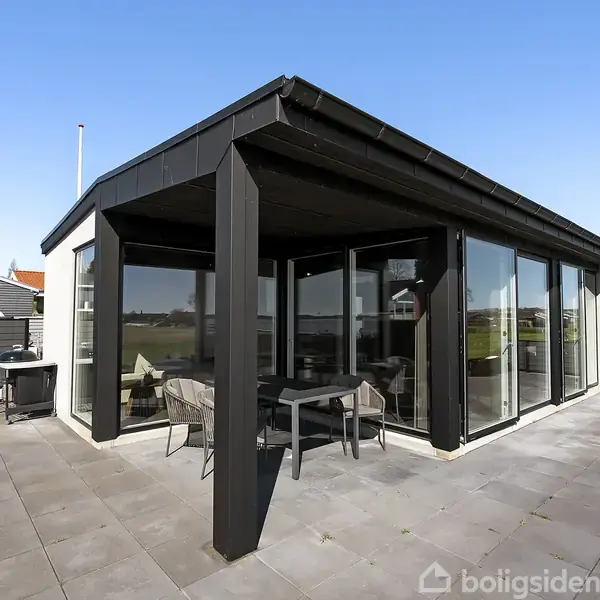 En moderne pavillon med glasvægge står på en stor flisebelagt terrasse. En grill og havemøbler ses ved siden af bygningen, og der er udsigt til et boligområde i baggrunden.