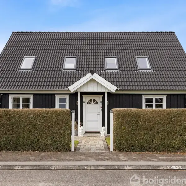 Et sort-hvidt hus med fire ovenlysvinduer står bag en lav hæk. Hoveddøren er centreret foran en flisebelagt sti, omgivet af en rolig villavej.