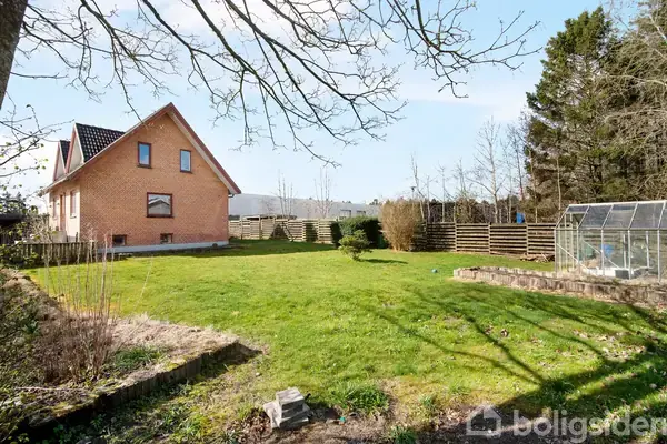 Et murstenshus med sort tag står i en have med grøn græsplæne. Der er en glasdrivhus til højre, omkranset af et træhegn og træer.