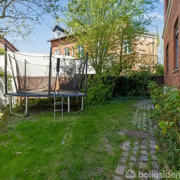 Trampolin står i en grøn have, omringet af hække og et murstenshus på højre side. Plankeværk og boligblokke ses i baggrunden under en blå himmel.