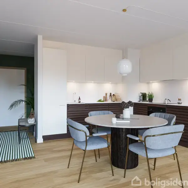 En rund spisebord med blå stole står i et moderne køkken. Køkkenet har hvide og mørkebrune skabe, hængende lamper, og planter som dekoration. Gulvet er lyst træ.