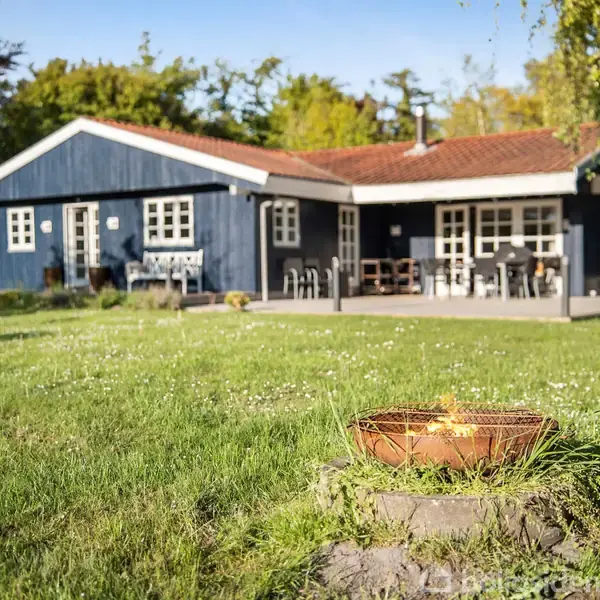 Træhus med blå facade og rødt tag står stille i en frodig have. Foran huset er der en græsplæne med en rund bålplads midt i det grønne miljø.
