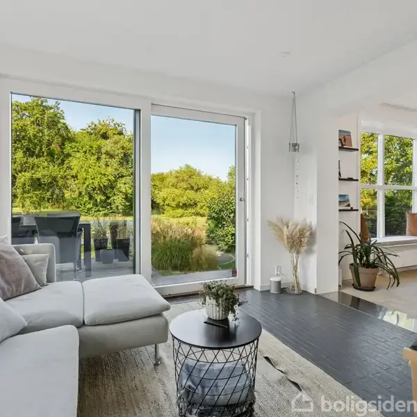 En lys stue med en hvid sofa vender mod en stor glasdør, der åbner ud til en terrasse med udsigt til grønne træer. En sort rund bord med planter står foran sofaen. En åben reol og køkkenet ses i baggrunden.