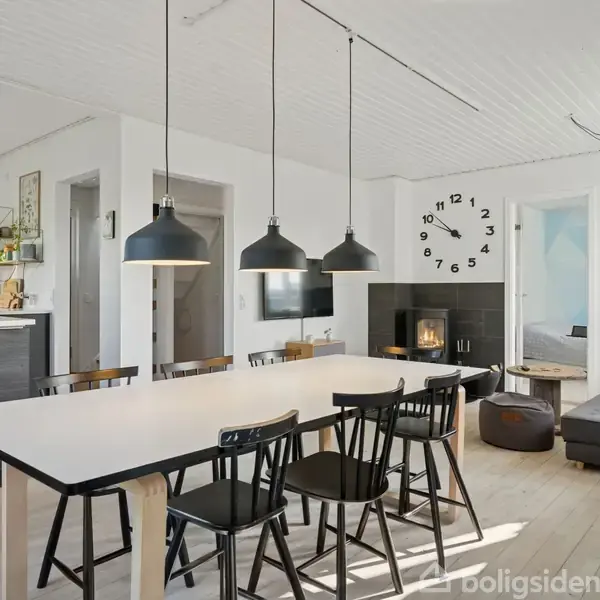 Et moderne køkken-alrum med spisebord omgivet af sorte stole. Tre sorte pendellamper hænger over bordet. I baggrunden er en grå sofa, en væg med ur og pejs.