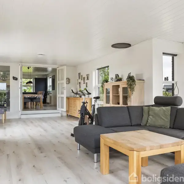Gråt sofaarrangement står i en lys stue med parketgulv, hvor et træbord står foran. I baggrunden ses et spiseområde med grønne planter og træmøbler.