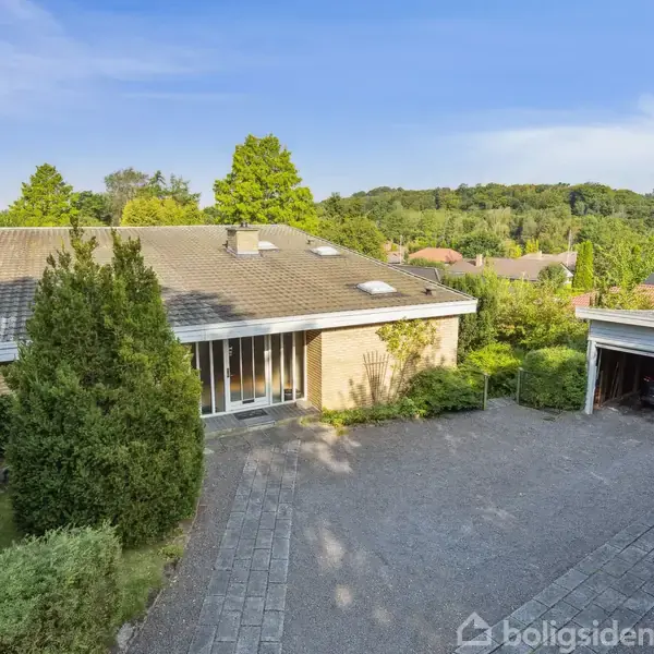 Hus med fladt tag står stille i en grøn have. I forgrunden grusindkørsel med tilhørende carport til højre. Omkringliggende træer og buske skaber en rolig atmosfære.