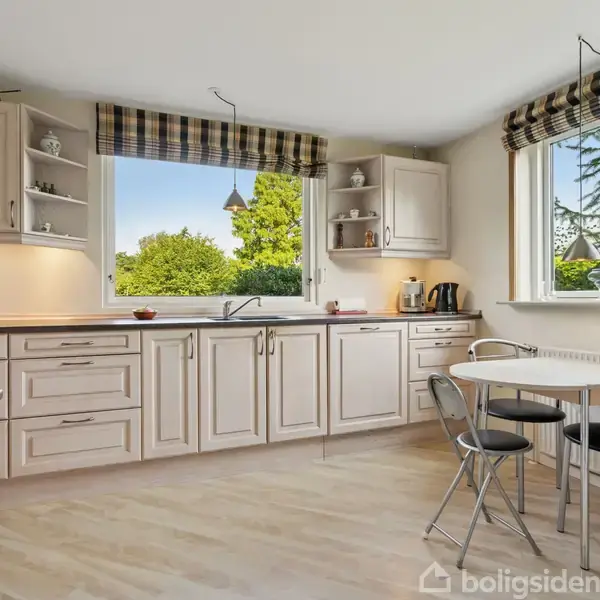 Et lyst køkken med hvide skabe og skuffer. En metalstole og -bord ved vinduet, som har udsigt til grønne træer. Kaffe- og teapparater står på bordet.