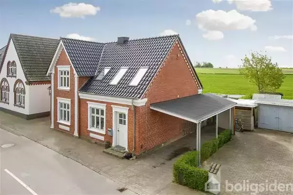 Rødt murstenshus med sort tag og tagvinduer, stående ved en vej. Huset har en overdækket indkørsel til højre, med en gårdsplads omgivet af grønne marker og nabobygninger i baggrunden.