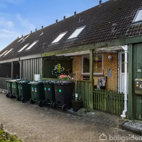 Rækkehus med grøn træfacade, række af skraldespande foran indgang. Vej med fortov og græs til venstre. Nummeret "35" er synligt på postkassen ved døren.