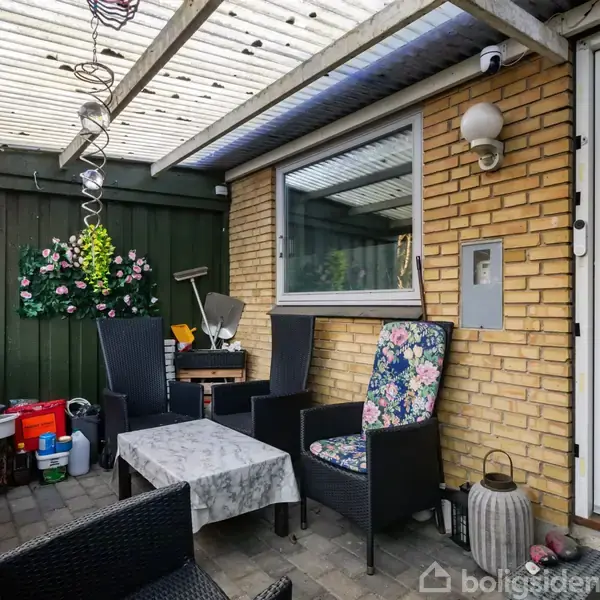 Terrasse med sorte stole og bord, beklædt med en dug, placeret foran en gul murstensvæg. Grønne planter hænger på væggen, og en glasdør ses til højre.