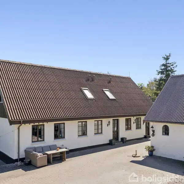 Gårdhus med hvid facade og mørkt tag, placeret i en gårdsplads. Sofa står langs muren, omgivet af en lys himmel og få træer i baggrunden.