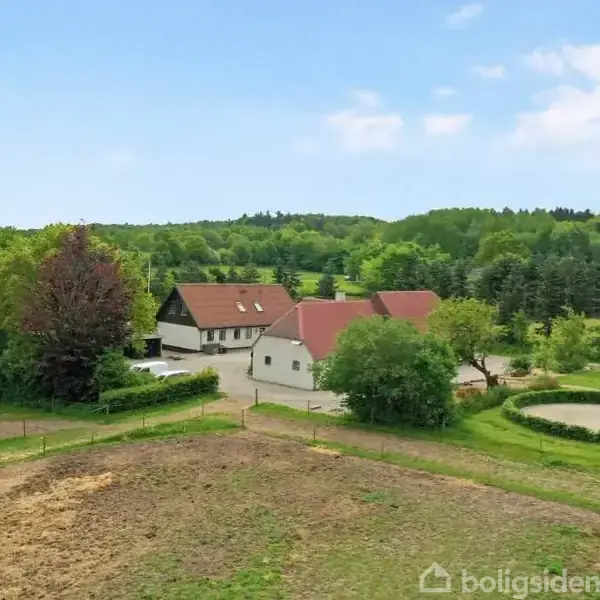 Gårdbygninger med rødbrune tage står omgivet af grønne træer og plæner. I forgrunden er en indhegnet græsmark og en cirkulær grusvej. Landligt område under blå himmel.