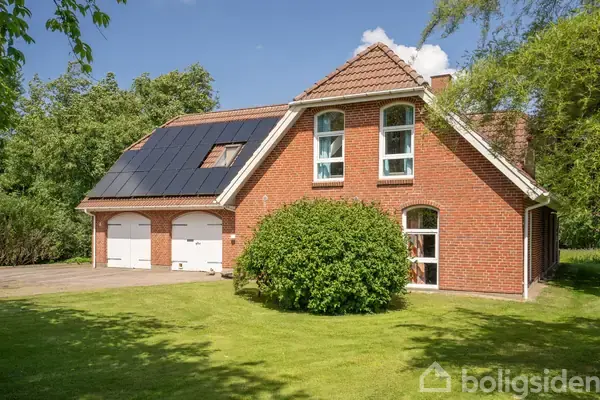 Rødt murstenshus med solpaneler på taget står på en velplejet græsplæne. To hvidmalede garageporte er synlige, og grønne buske omgiver huset i et solrigt område.