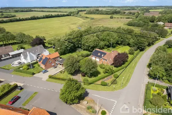 Et rødt murstenshus med solpaneler på taget står stille omgivet af en velholdt grøn græsplæne, store træer og buske. To hvide garageporte ses i venstre side.
