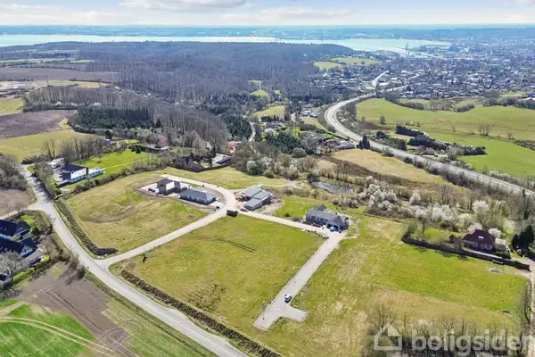 Landområde set fra luften med en vej, der snor sig gennem grønne marker, omgivet af skove. Husgrupper ligger spredt i landskabet, med byen og vandet i baggrunden.