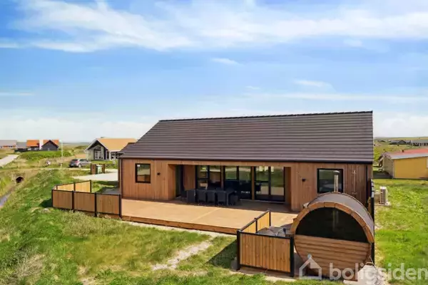 Et moderne træhus står på græsset i et kystområde. Huset har store vinduer og en terrasse med hegn samt en rund sauna foran.