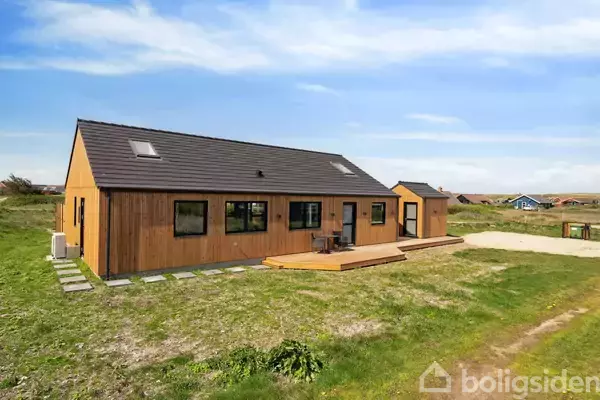 Et moderne træhus står stille på en græsplæne, omgivet af en blå himmel. Terrasse med borde og stole i forgrunden.