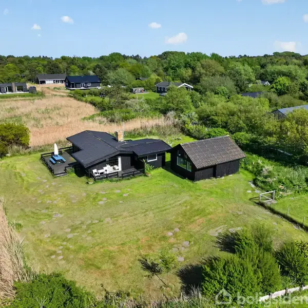 En sort træhytte med terrasse står stille midt på en stor græsplæne. Omgivet af frodig vegetation og andre små bygninger i en landlig, grøn landskabskontekst.