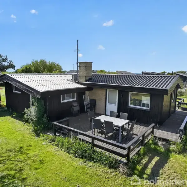 Et sort sommerhus med en terrasse, hvor der står et bord og fire stole. Det ligger i en grøn have med træer og en blå himmel i baggrunden.
