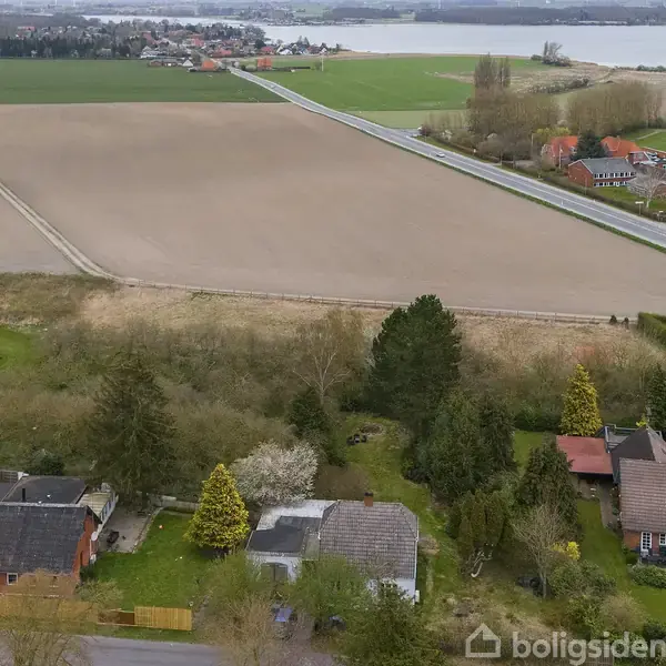 Huse ligger side om side i et grønt landdistrikt; veje skærer gennem marker mod en fjord i baggrunden, omkranset af spredte træer og små bygninger.