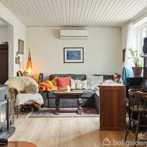 Stue med sofa og sofabord i midten, omgivet af lænestol, klaver og brændeovn. På væggen hænger et billede. Vindue og planter til højre. Løbelys på bordpladen.