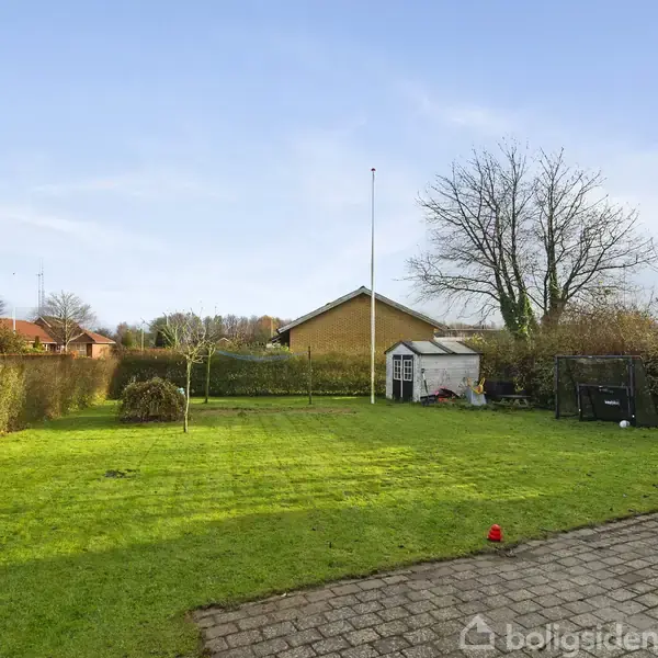 Græsplæne med flagstang står i en velplejet have, omgivet af hække og træer, med en rød murstensbygning til højre og en gul bygning i baggrunden under en blå himmel.