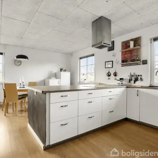 Et moderne køkken med hvide skabe og skuffer, ovenpå en lys træbordplade. I baggrunden står et spisebord med stole nær vinduer, der lukker naturligt lys ind.