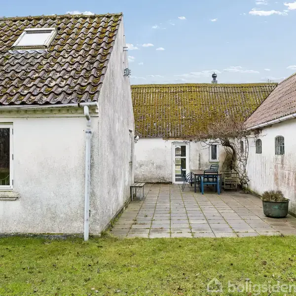 Hus med hvid facade og rødt tegltag står stille, flankeret af en flisebelagt gårdsplads. Et bord med stole står i gården mellem huset og en tilstødende bygning. Græsplæne omringer området.