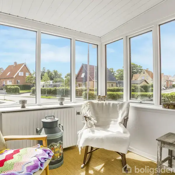 Stol står stille på solrig veranda, omgivet af store vinduer, der åbner op til en udsigt over et boligkvarter med huse. Et farverigt tæppe ligger på en anden stol.
