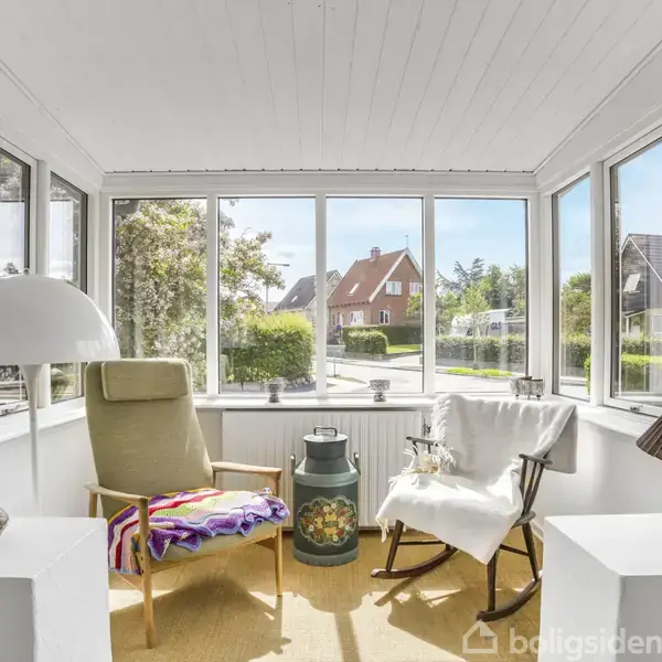 En lys udestue med to stole, hvoraf den ene har et farverigt tæppe. En lampe og pyntegenstande står i rummet, omgivet af store vinduer mod en grøn have og huse.