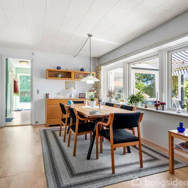 Spisebord med seks stole står på et tæppe i en spisestue. Rummet er lyst med store vinduer, der viser en grøn have udenfor. En lampe hænger over bordet.