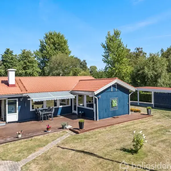 Blå træbungalow med rødt tag står på en velplejet græsplæne med en træterrasse. Terrassen har havemøbler og en markise. Omgivet af træer og buske i en fredelig have.