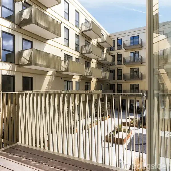 Flere altaner strækker sig ud fra facaden på en moderne boligblok. Bygningen omkranser en lys gårdhave med planter og siddepladser.