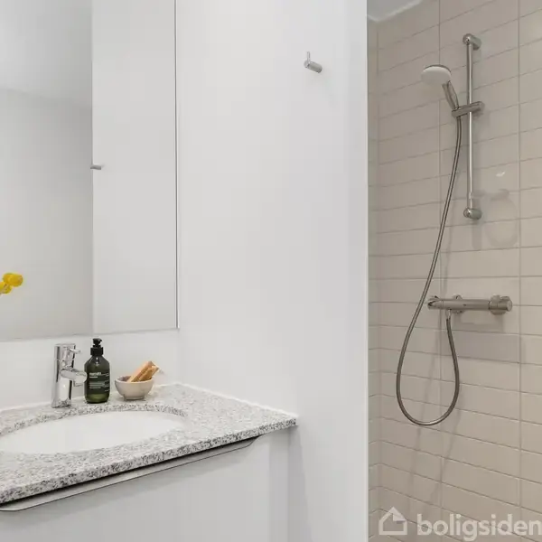 En moderne badeværelseshåndvask med en spejlskab over, omgivet af hvide vægge. Ved siden af ses en bruser med flisebelagt væg. På bordet står en vase med gule blomster og en sæbedispenser.