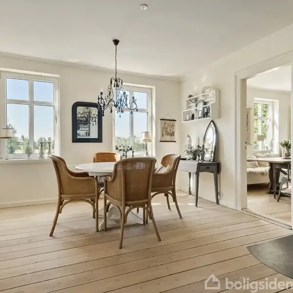 Et spisebord med stole står i et lyst rum. En elegant lysekrone hænger over bordet. Til højre er en pejs, og der er adgang til et tilstødende rum. Ingen synlig tekst.