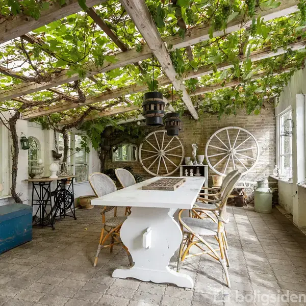 Et stort hvidt bord med stole står i et lyst rum med store vinduer. Vinstokke hænger fra loftsbjælker, og der er dekorative hjul på bagvæggen.
