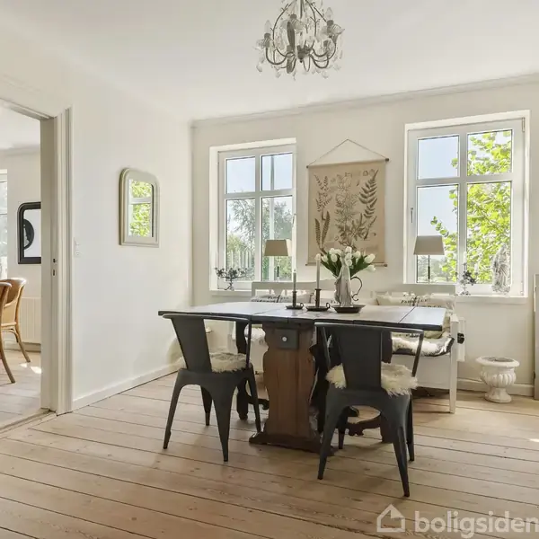 Spisebord med stole, dekoreret med vase og blomster, står i et lyst rum med trægulv og hvidmalede vægge. Store vinduer giver udsigt til grønne træer udenfor.